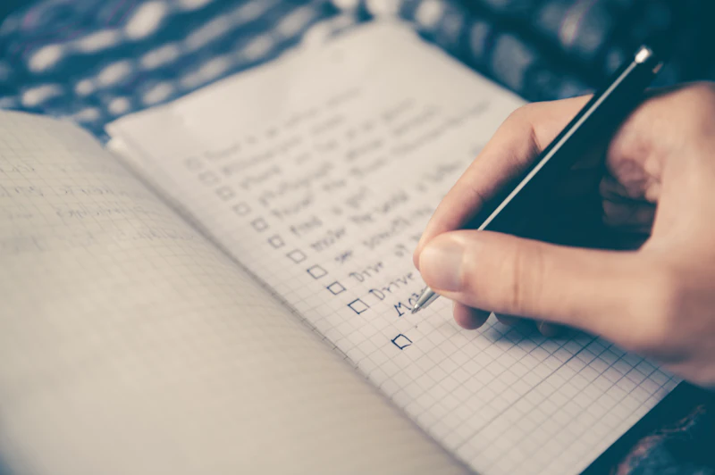 A hand ticking off a handwritten checklist in a grid-paper notebook.