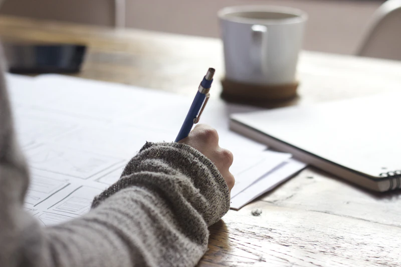 A person in a gray sweater writes notes on paper at a wooden table, a coffee cup beside them.