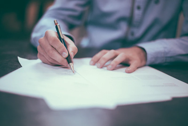 A person in a collared shirt signs a printed document at a desk.